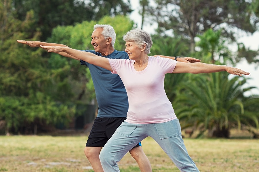 An elderly man and woman doing a yoga pose side by side outdoors