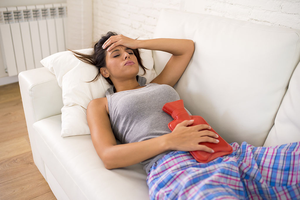 A Woman Lying On a Couch In Pain Holding a Heating Pad To Her Stomach Adenomyosis Treatments