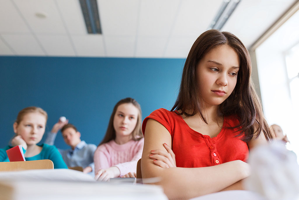 Closeup of a Teenage Girl Sitting at a Desk Sad With Two Other Girls Gossiping Behind Her Primary Amenorrhea